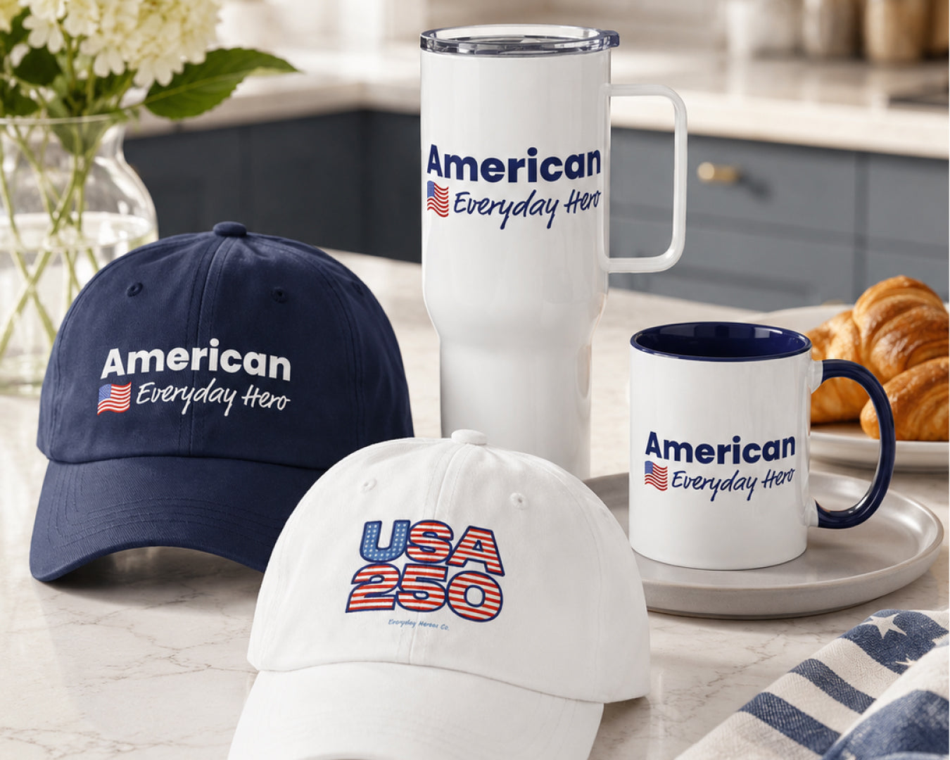 A coordinated set of branded merchandise on a kitchen counter: a navy baseball cap, a white baseball cap with a large USA 250 graphic, a tall white handled travel mug, and a white mug with a navy rim and handle, all printed with the slogan 'American Everyday Hero' and a small American flag. A croissant and flowers are visible in the background.
