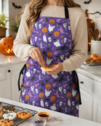 A woman wearing a purple Halloween apron with a ghost, pumpkin, bat, and witch pattern, standing in a kitchen and sprinkling icing on small cookies.