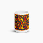 The side of a white ceramic mug with a colorful autumn pattern, featuring red, orange, and yellow maple leaves on a dark background with decorative leaf borders photographed against a light-grey studio backdrop.