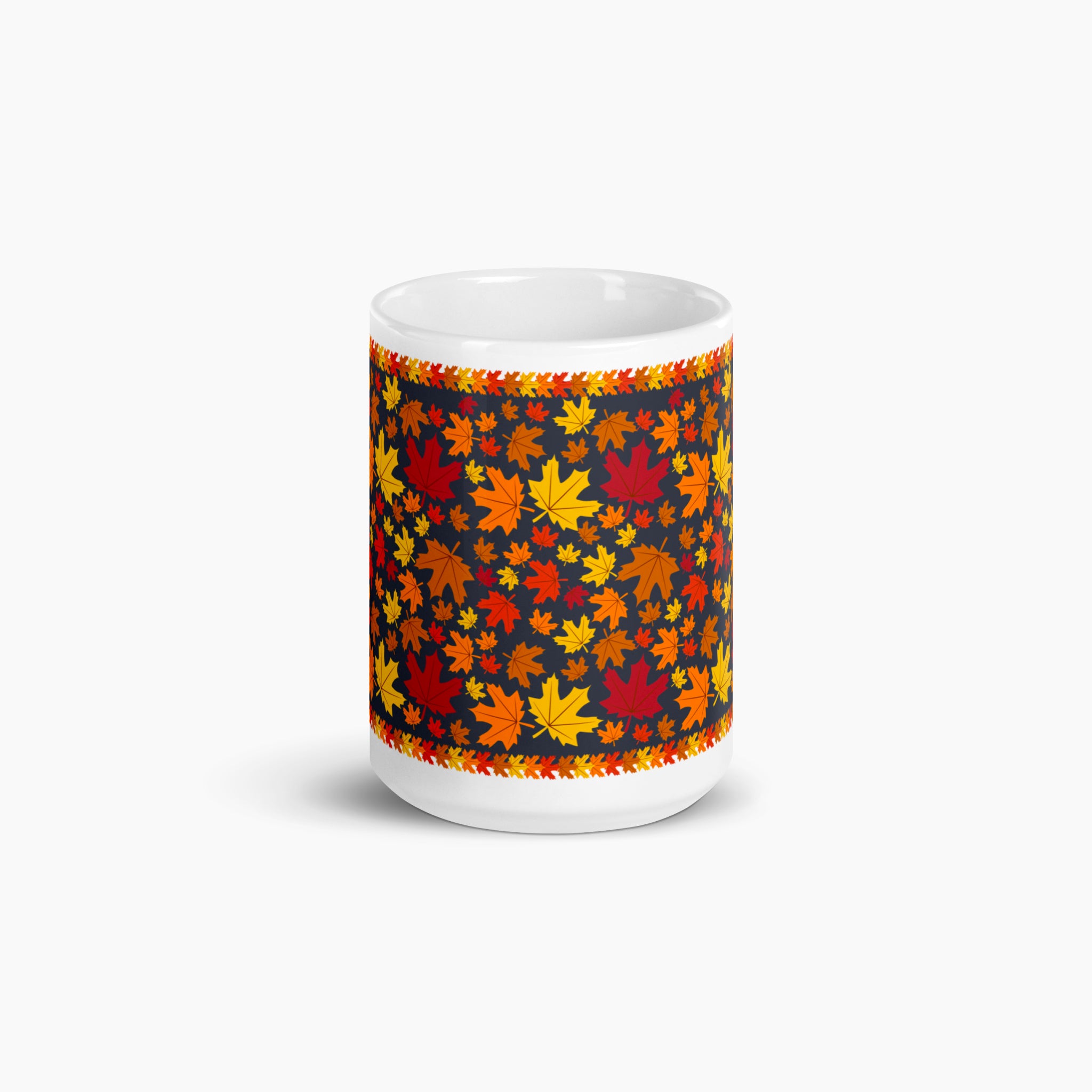 The side of a white ceramic mug with a colorful autumn pattern, featuring red, orange, and yellow maple leaves on a dark background with decorative leaf borders photographed against a light-grey studio backdrop.