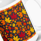 A close-up of a white ceramic mug featuring a wrap-around design of colorful autumn maple leaves in shades of orange, yellow, red, and brown on a dark navy background. Photographed against a light-grey studio backdrop.