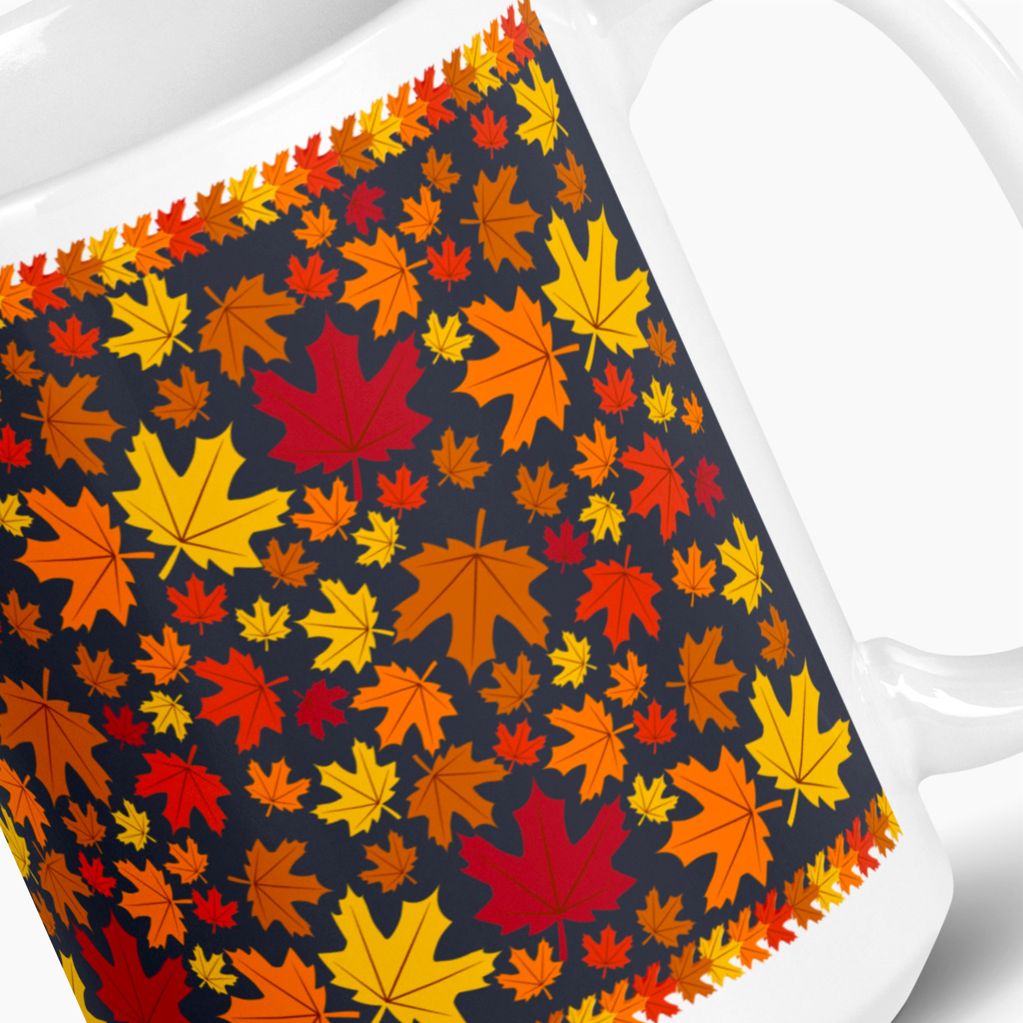 A close-up of a white ceramic mug featuring a wrap-around design of colorful autumn maple leaves in shades of orange, yellow, red, and brown on a dark navy background. Photographed against a light-grey studio backdrop.