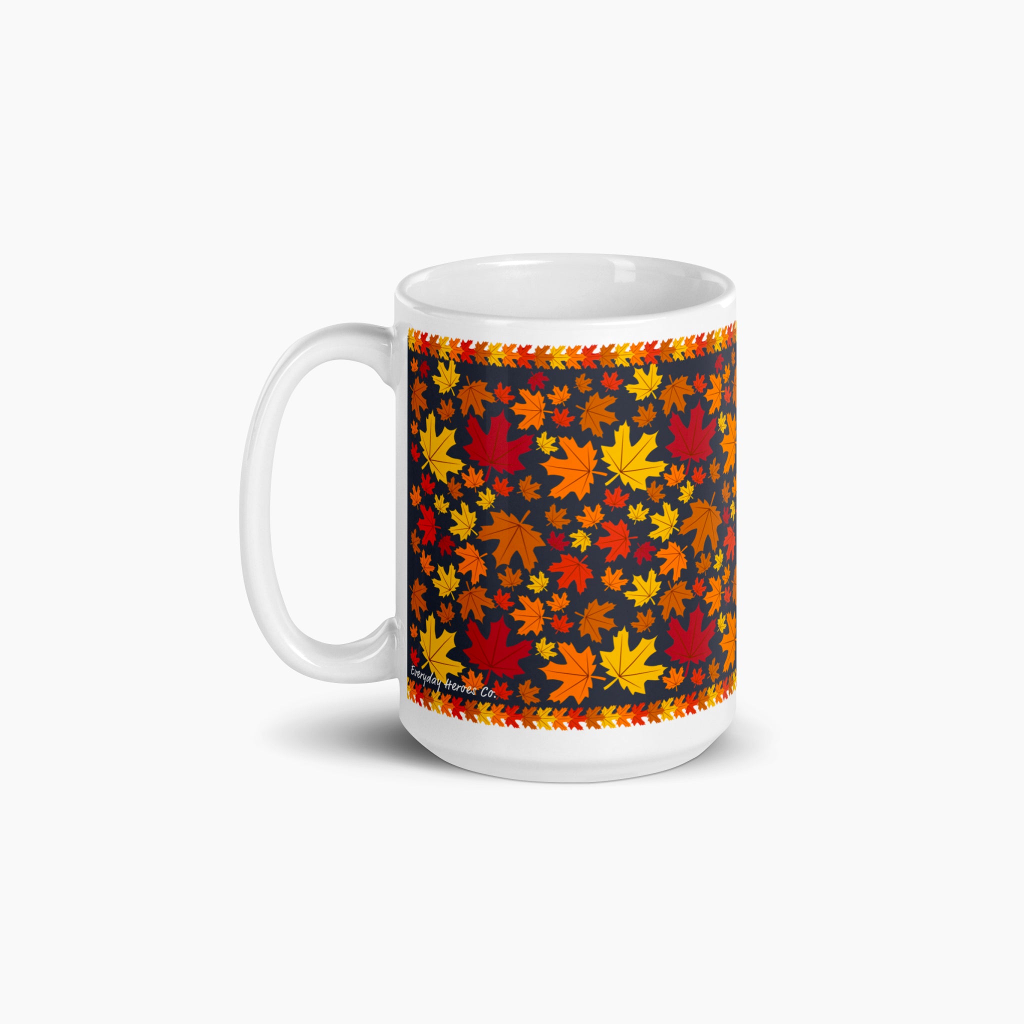 The back side of a white ceramic mug with a colorful autumn pattern, featuring red, orange, and yellow maple leaves on a dark background with decorative leaf borders photographed against a light-grey studio backdrop.