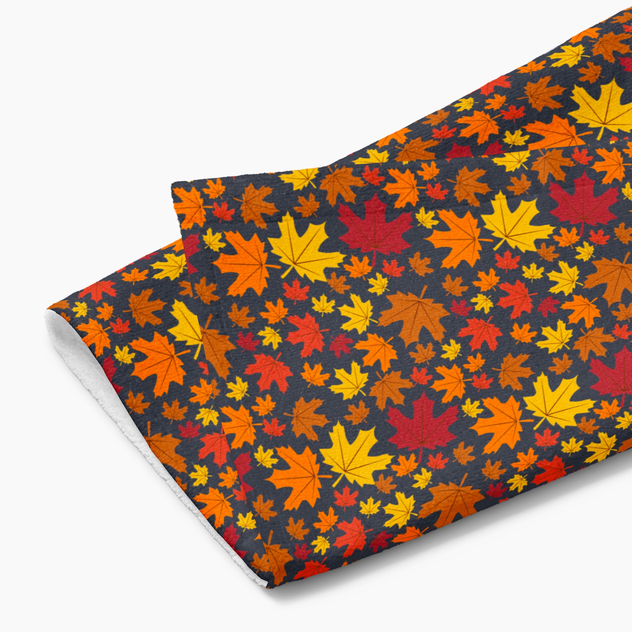 A folded rectangular fabric kitchen towel featuring an all-over autumn maple leaf pattern in red, orange, yellow, and brown on a dark charcoal background, photographed against a light gray backdrop.