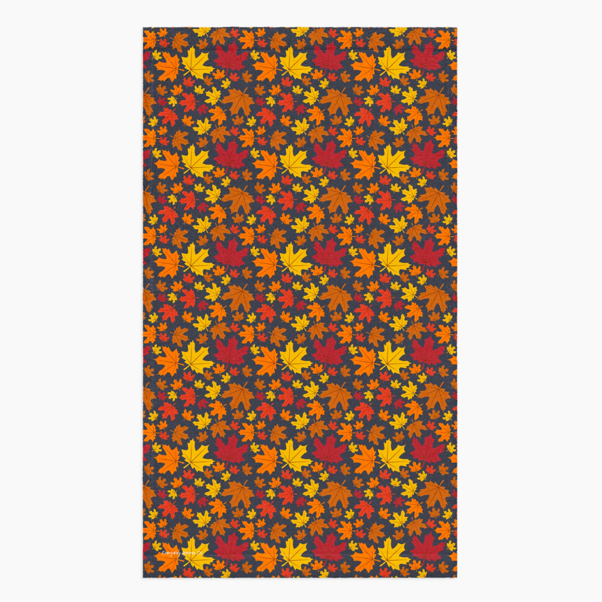 A rectangular fabric kitchen towel featuring an all-over autumn maple leaf pattern in red, orange, yellow, and brown on a dark charcoal background, shown flat against a light gray backdrop.