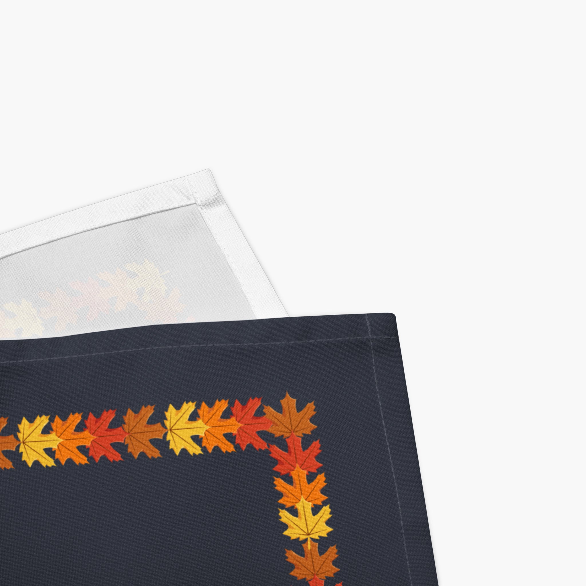 Close-up of a folded cloth napkins with a colorful autumn leaf border pattern in orange, yellow, and red, staged on a light gray background.