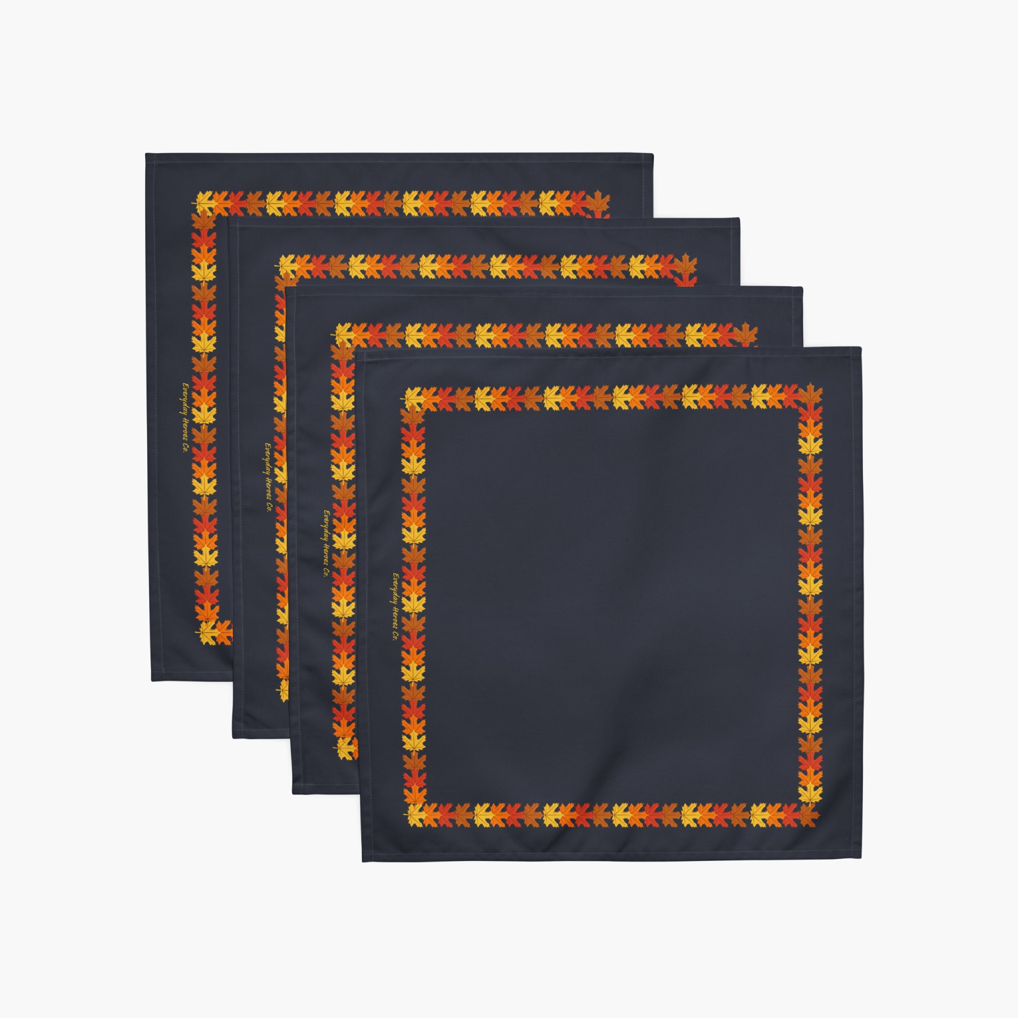 Four flat dark navy cloth napkins with a colorful autumn leaf border pattern in orange, yellow, and red, staggered on a light gray background.