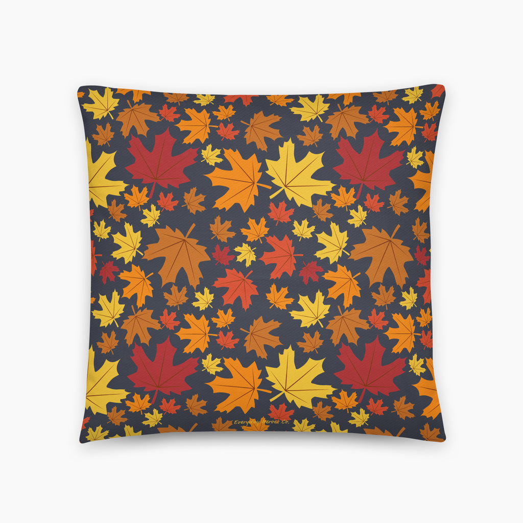 A square throw pillow featuring a dense pattern of colorful autumn maple leaves in shades of red, orange, yellow, and brown on a dark charcoal gray background, shown against a plain light grey studio backdrop.