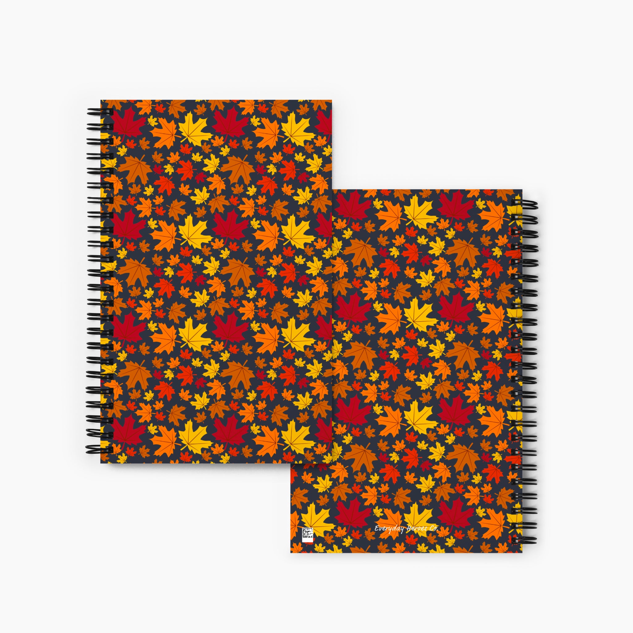 Front and back cover spiral-bound notebook featuring pattern of autumn maple leaves in red, orange, yellow, and brown on a dark background, shown against a light gray studio backdrop.