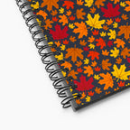 A spiral-bound notebook shown at an angle on a light-grey backdrop, featuring a dark charcoal cover decorated with a colorful pattern of autumn maple leaves in red, orange, yellow, and brown.