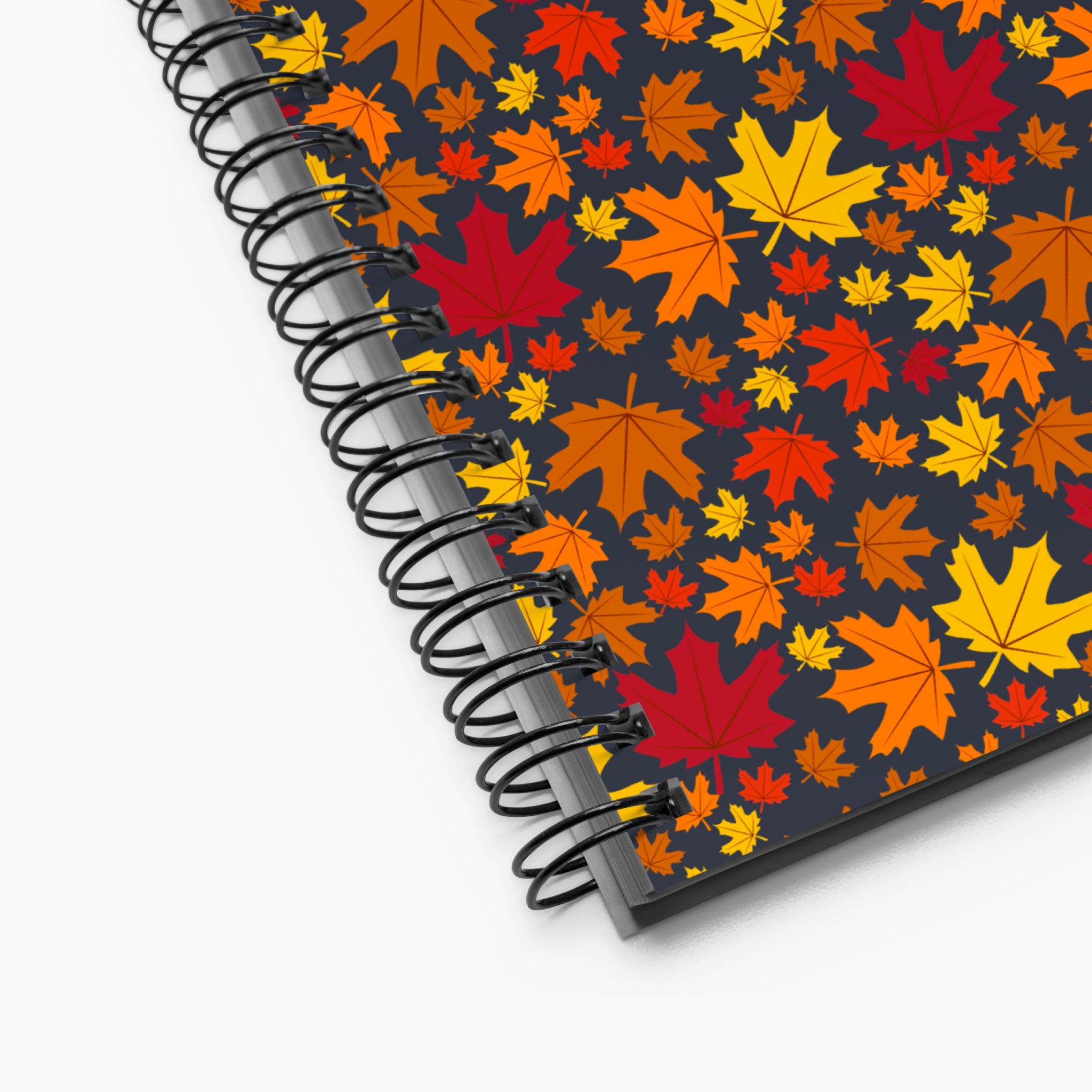 A spiral-bound notebook shown at an angle on a light-grey backdrop, featuring a dark charcoal cover decorated with a colorful pattern of autumn maple leaves in red, orange, yellow, and brown.