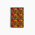 A spiral-bound notebook with a cover pattern of overlapping autumn maple leaves in red, orange, yellow, and brown on a dark background, shown against a light gray studio backdrop.