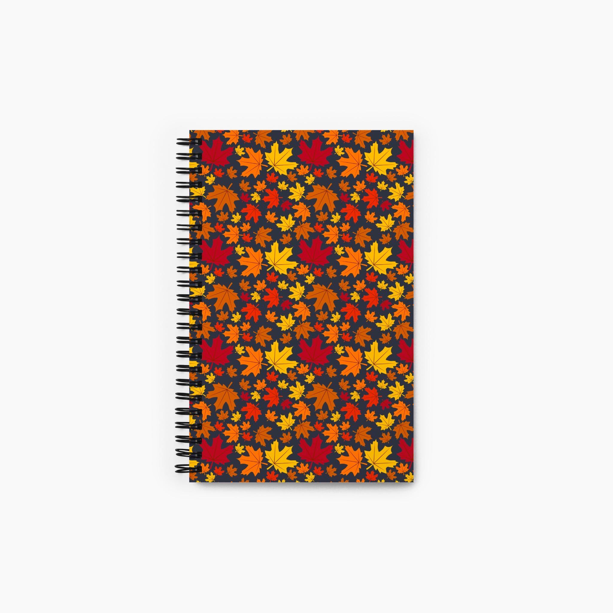 A spiral-bound notebook with a cover pattern of overlapping autumn maple leaves in red, orange, yellow, and brown on a dark background, shown against a light gray studio backdrop.