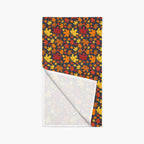 A folded table runner with a dark background covered in a dense pattern of red, orange, yellow, and brown autumn maple leaves. The bottom corner is folded over to show the white reverse side with a lighter pastel leaf pattern.