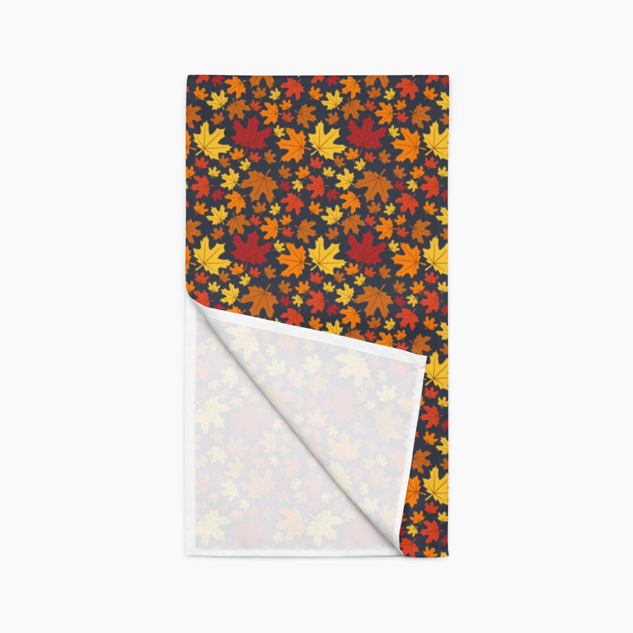 A folded table runner with a dark background covered in a dense pattern of red, orange, yellow, and brown autumn maple leaves. The bottom corner is folded over to show the white reverse side with a lighter pastel leaf pattern.