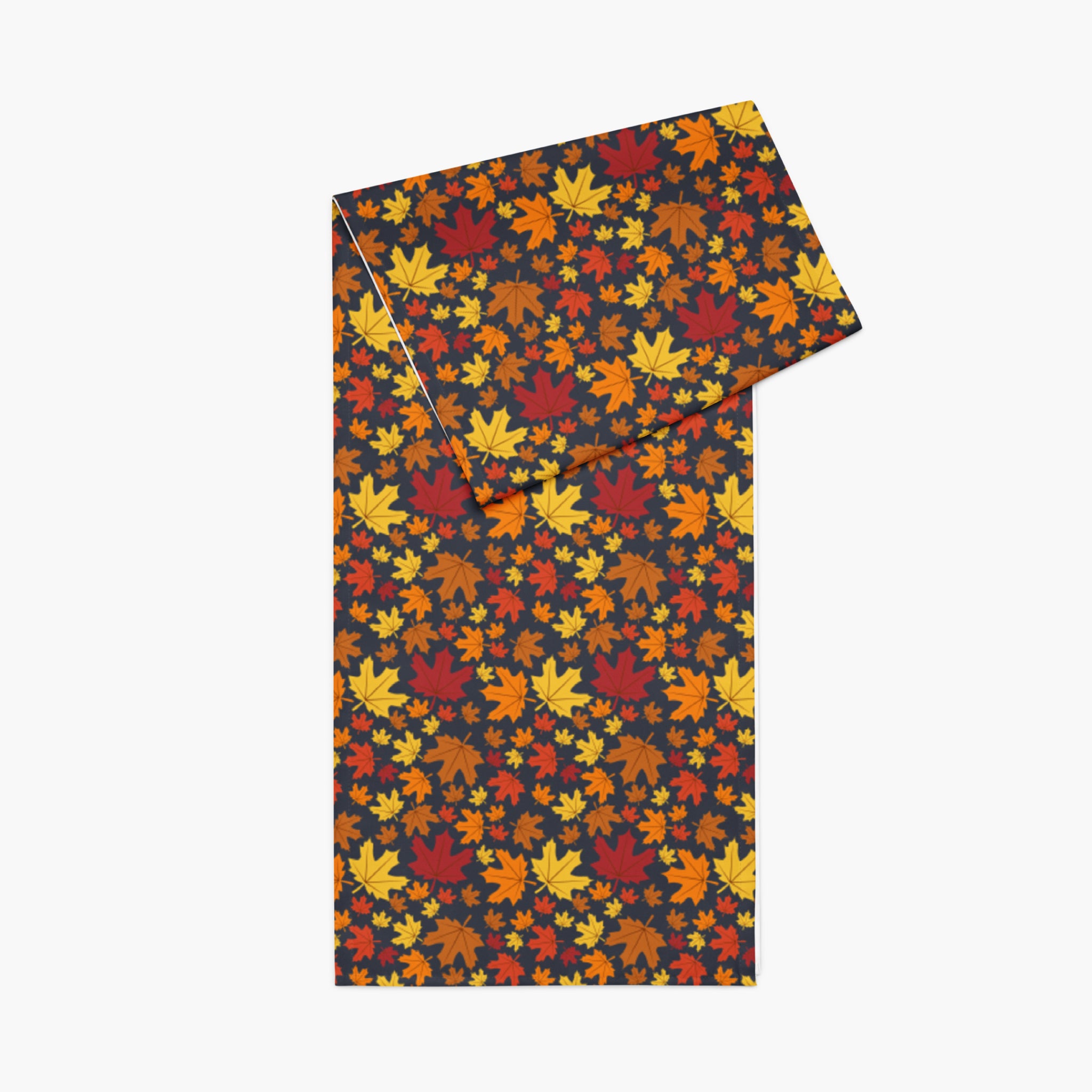 A folded rectangular table runner, featuring an all-over autumn maple leaf pattern in red, orange, yellow, and brown on a dark charcoal background, shown on a plain light gray background.