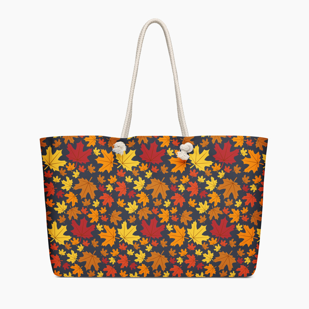 A tote bag with a dense autumn maple leaf pattern in red, orange, yellow, and brown on a dark charcoal background, featuring  white handles.
