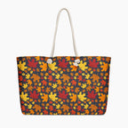 A tote bag with a dense autumn maple leaf pattern in red, orange, yellow, and brown on a dark charcoal background, featuring  white handles.