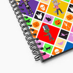 A close-up of a spiral-bound notebook showing a colorful Halloween-themed cover design with cartoon skeletons, skulls, ghosts, pumpkins, a green monster, bats, and black cats on a light grey background.