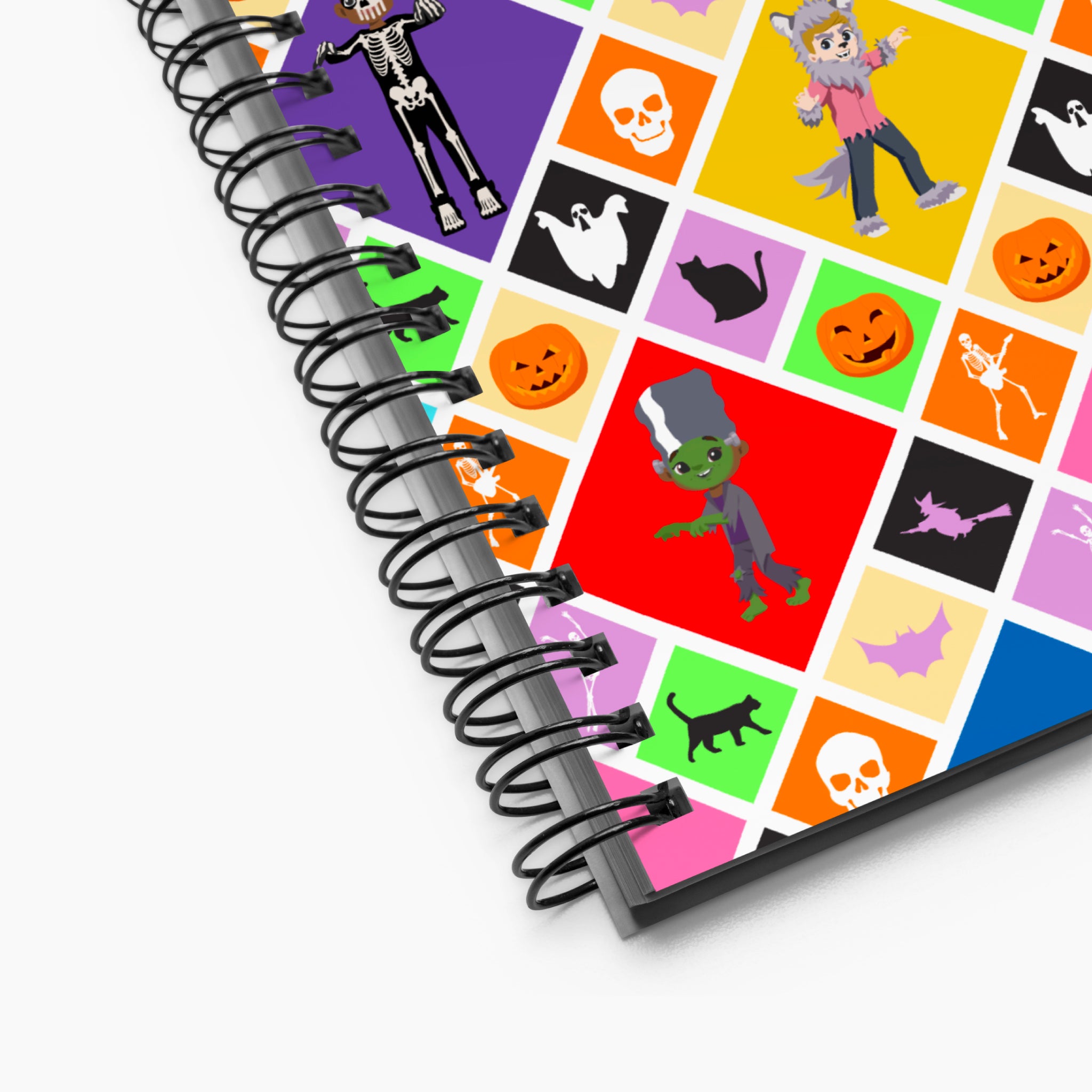 A close-up of a spiral-bound notebook showing a colorful Halloween-themed cover design with cartoon skeletons, skulls, ghosts, pumpkins, a green monster, bats, and black cats on a light grey background.