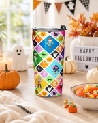 A tall insulated tumbler with a clear lid and black flip top stands on a wooden table in a Halloween-themed setting. The tumbler has a colorful diamond-patterned wrap featuring pumpkins, ghosts, witches, and other spooky motifs.