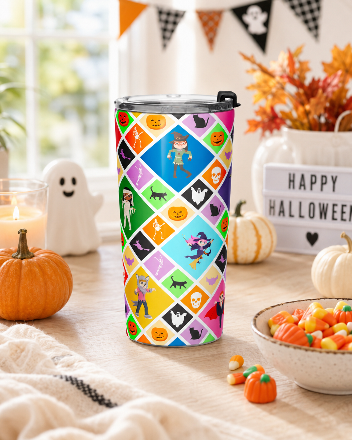 A tall insulated tumbler with a clear lid and black flip top stands on a wooden table in a Halloween-themed setting. The tumbler has a colorful diamond-patterned wrap featuring pumpkins, ghosts, witches, and other spooky motifs.