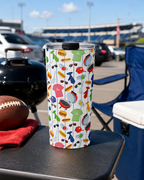 Colorful tumbler with a Dad-themed pattern on a table outdoors, with cars and a chair in the background at a tailgate.