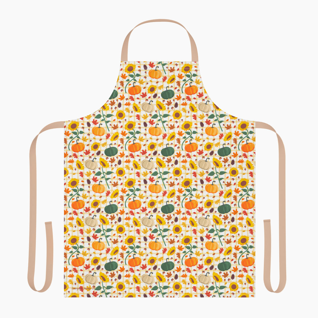 A white apron with a beige neck loop and waist ties, featuring an all-over autumn pattern of pumpkins, sunflowers, gourds, and fall leaves.