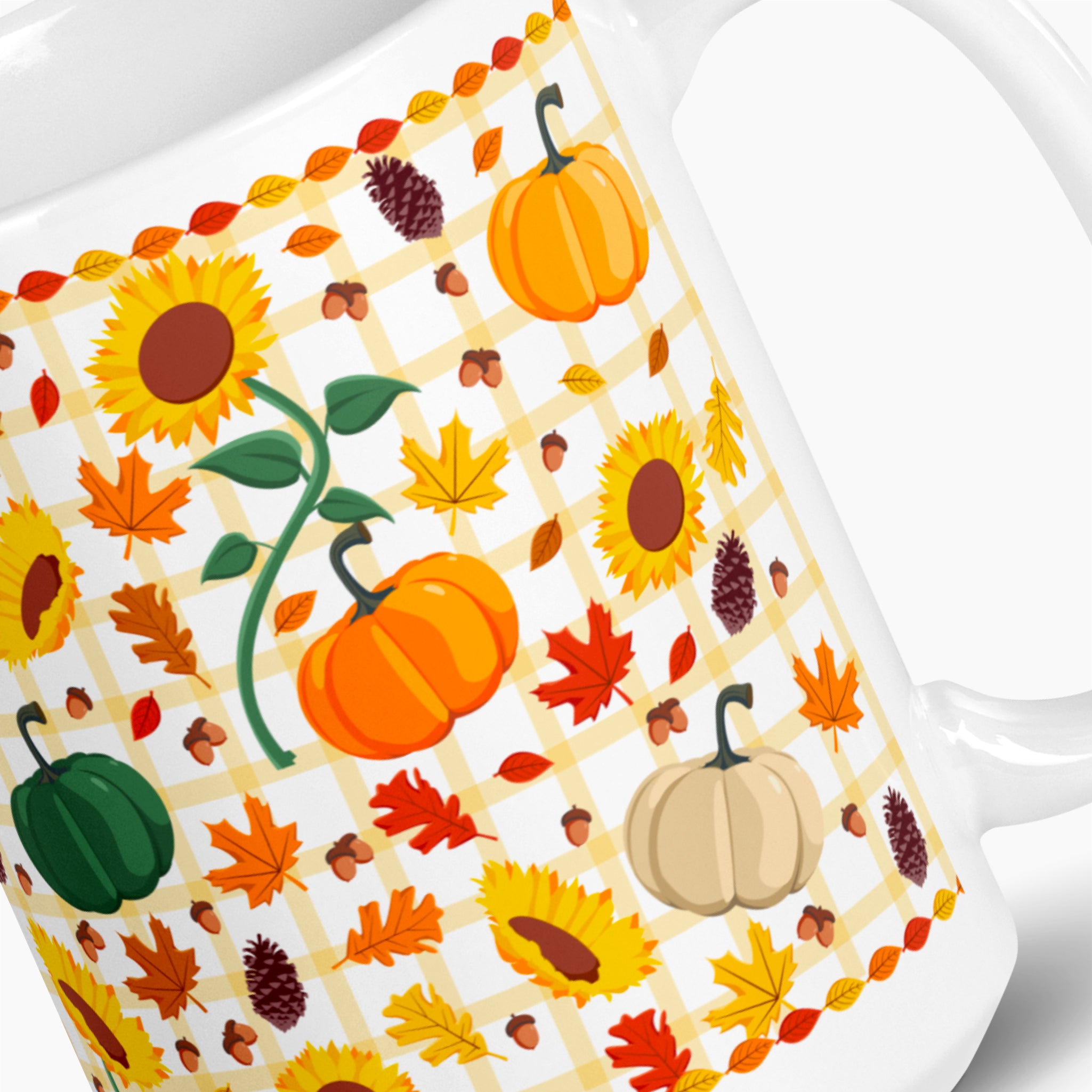 A close-up of a white ceramic mug decorated with an autumn pattern featuring pumpkins, sunflowers, maple leaves, acorns, pinecones, and a yellow-and-white gingham grid.