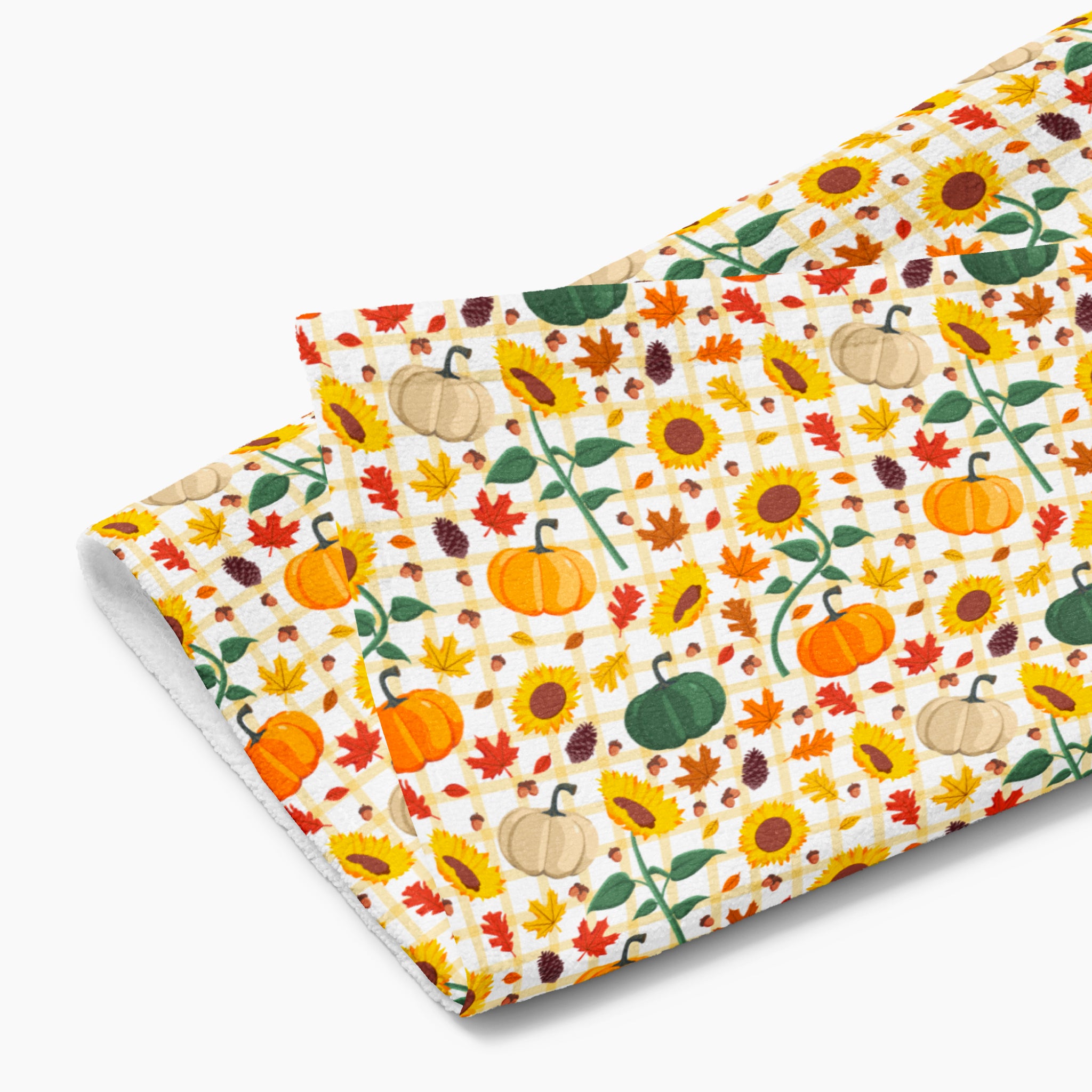 A folded dish towel featuring a colorful autumn pattern with pumpkins, sunflowers, maple leaves, pinecones, and plaid checks on a light-grey studio background.
