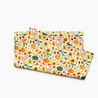 A folded fabric dish towel featuring a colorful autumn pattern with pumpkins, sunflowers, leaves, and acorns on a light-grey studio background.
