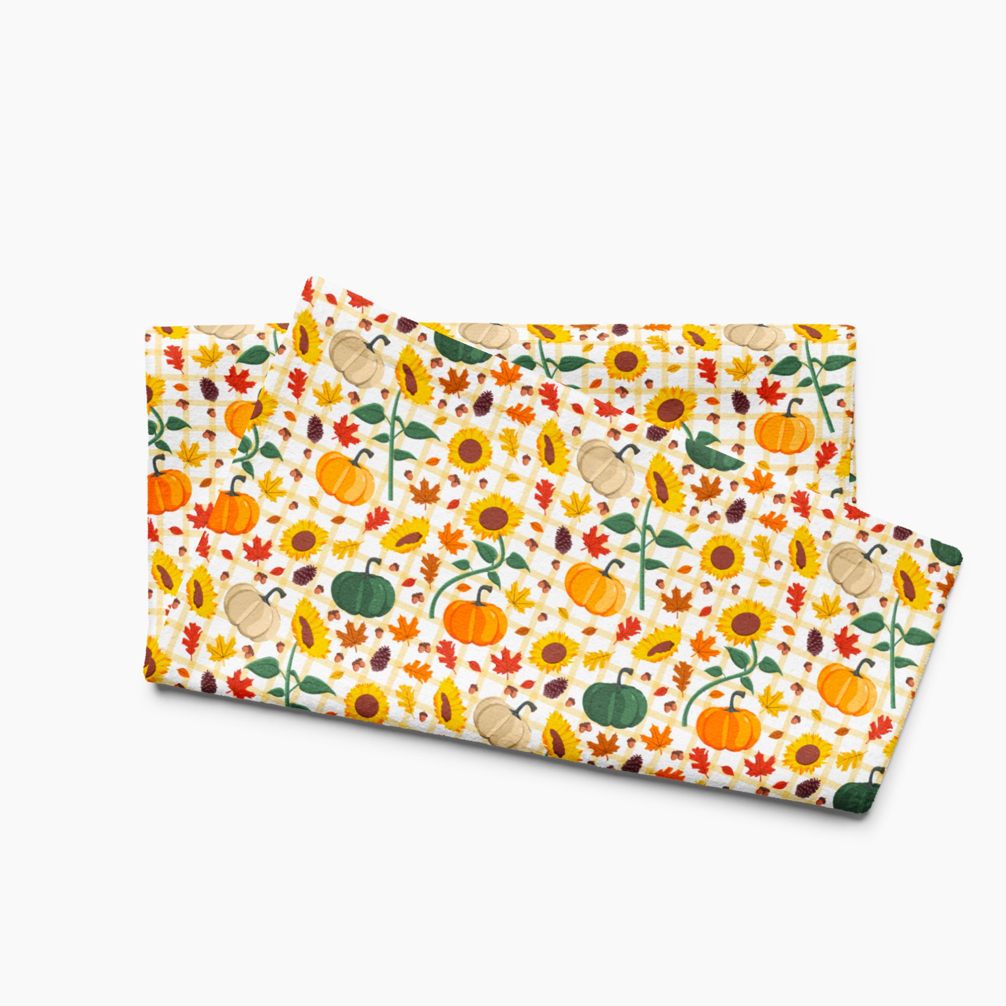 A folded fabric dish towel featuring a colorful autumn pattern with pumpkins, sunflowers, leaves, and acorns on a light-grey studio background.