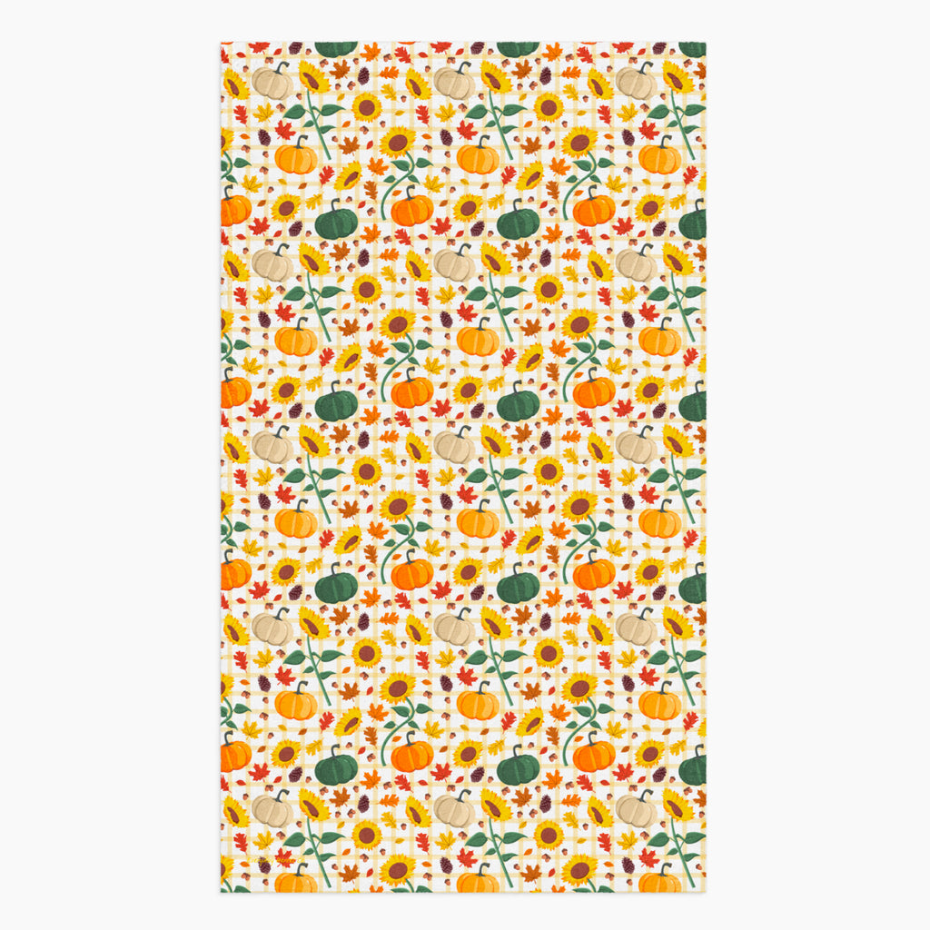 A dish towel featuring a repeating autumn pattern of pumpkins, sunflowers, gourds, acorns, and leaves on a light-grey studio backdrop.