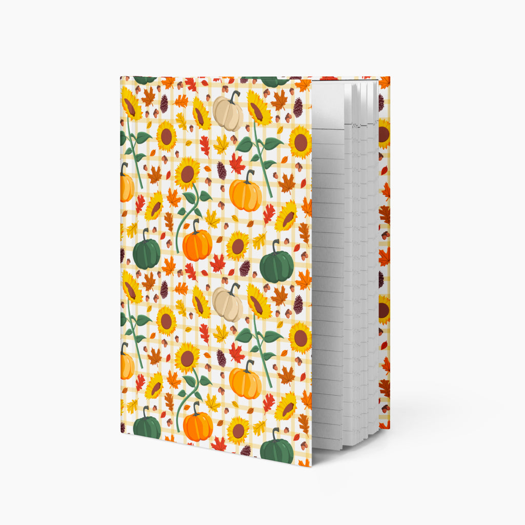 A hardcover notebook with a white cover decorated with a colorful autumn pattern of pumpkins, sunflowers, and leaves, standing upright on a light gray background.