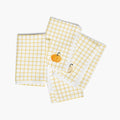 Four folded white cloth napkins with a yellow gingham check pattern, featuring a small pumpkin design on the corner of each napkin, displayed on a light-grey studio background.