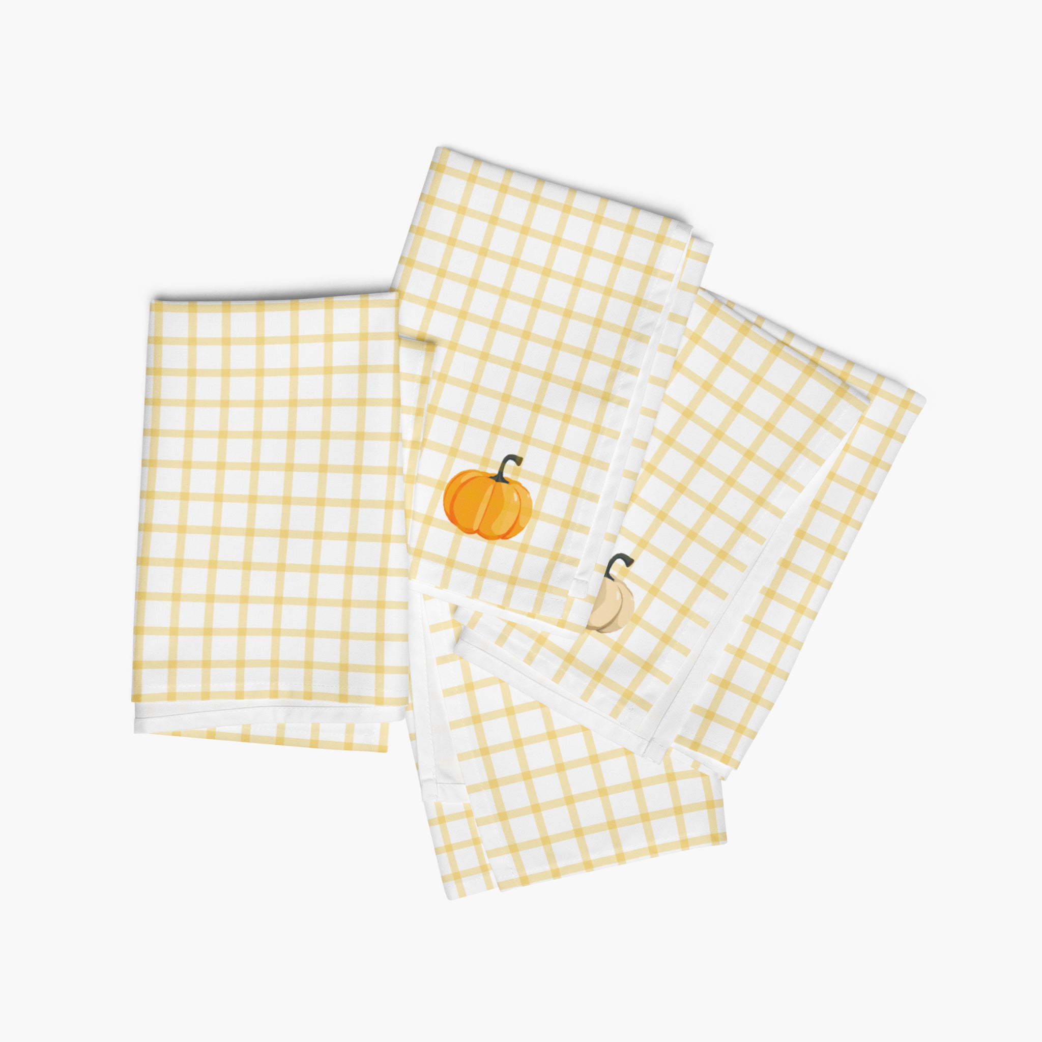 Four folded white cloth napkins with a yellow gingham check pattern, featuring a small pumpkin design on the corner of each napkin, displayed on a light-grey studio background.