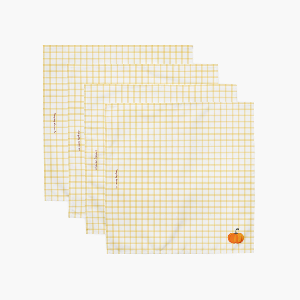 Four square white napkins with a yellow gingham check pattern, arranged in a staggered stack. Each napkin features a small pumpkin in the bottom right corner, and the napkins have subtle red text saying "Everyday Heroes Co" printed vertically along the left side.