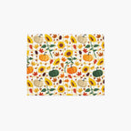 A rectangular placemat, featuring a repeating autumn pattern of pumpkins, sunflowers, maple leaves, and pinecones on a light-grey studio background.
