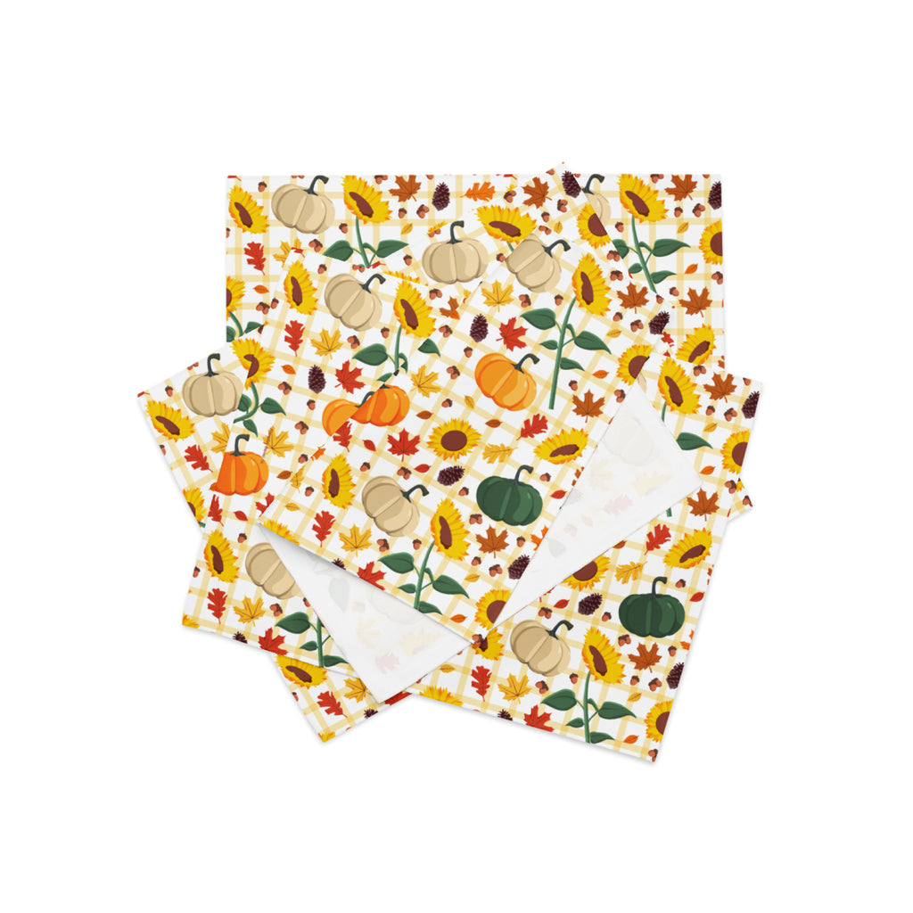 A stack of cloth placemats featuring a colorful autumn pattern with pumpkins, sunflowers, leaves, and acorns on a cream background, with one napkin showing a plain white reverse side.