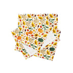 A stack of cloth placemats featuring a colorful autumn pattern with pumpkins, sunflowers, leaves, and acorns on a cream background, with one napkin showing a plain white reverse side.