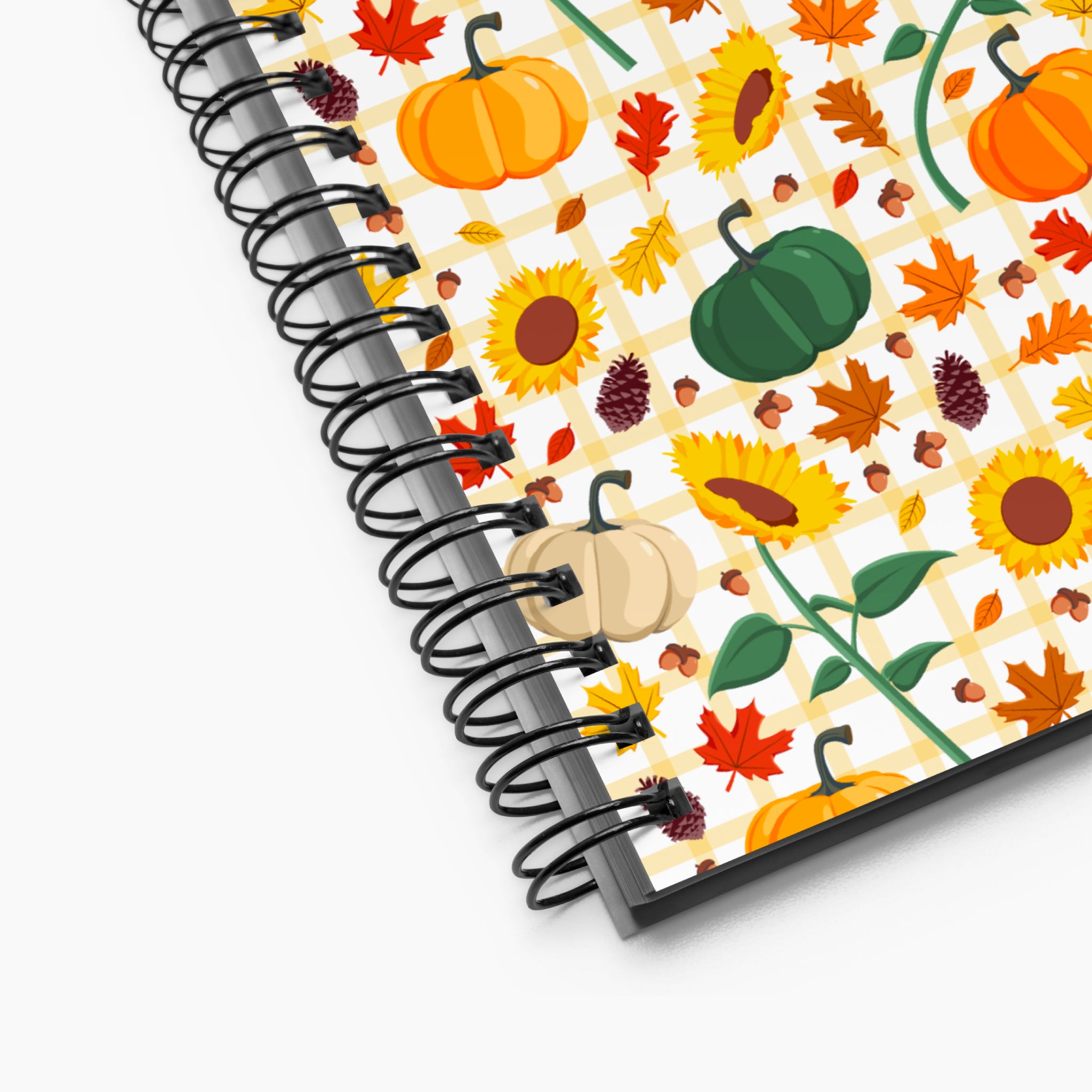 A spiral-bound notebook shown at an angle on a light-grey background, featuring a colorful autumn pattern with sunflowers, pumpkins, maple leaves, acorns, and pinecones over a light-yellow gingham pattern.