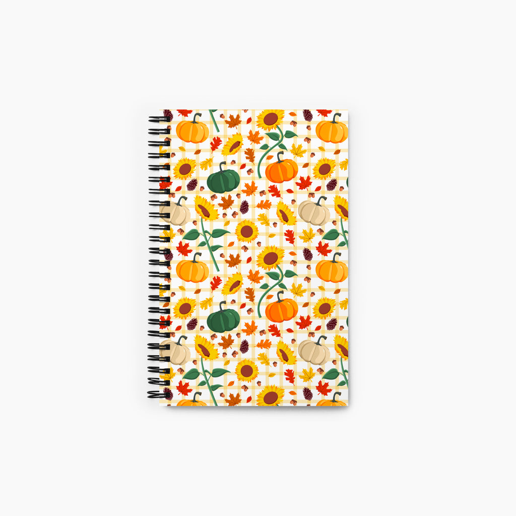 A spiral-bound notebook with a white cover decorated with an autumn pattern of pumpkins, sunflowers, maple leaves, pinecones, and acorns, shown against a plain light-grey background.