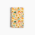 A spiral-bound notebook with a white cover decorated with an autumn pattern of pumpkins, sunflowers, maple leaves, pinecones, and acorns, shown against a plain light-grey background.