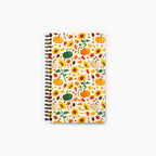 A spiral-bound notebook with a white cover decorated with an autumn pattern of pumpkins, sunflowers, maple leaves, pinecones, and acorns, shown against a plain light-grey background.