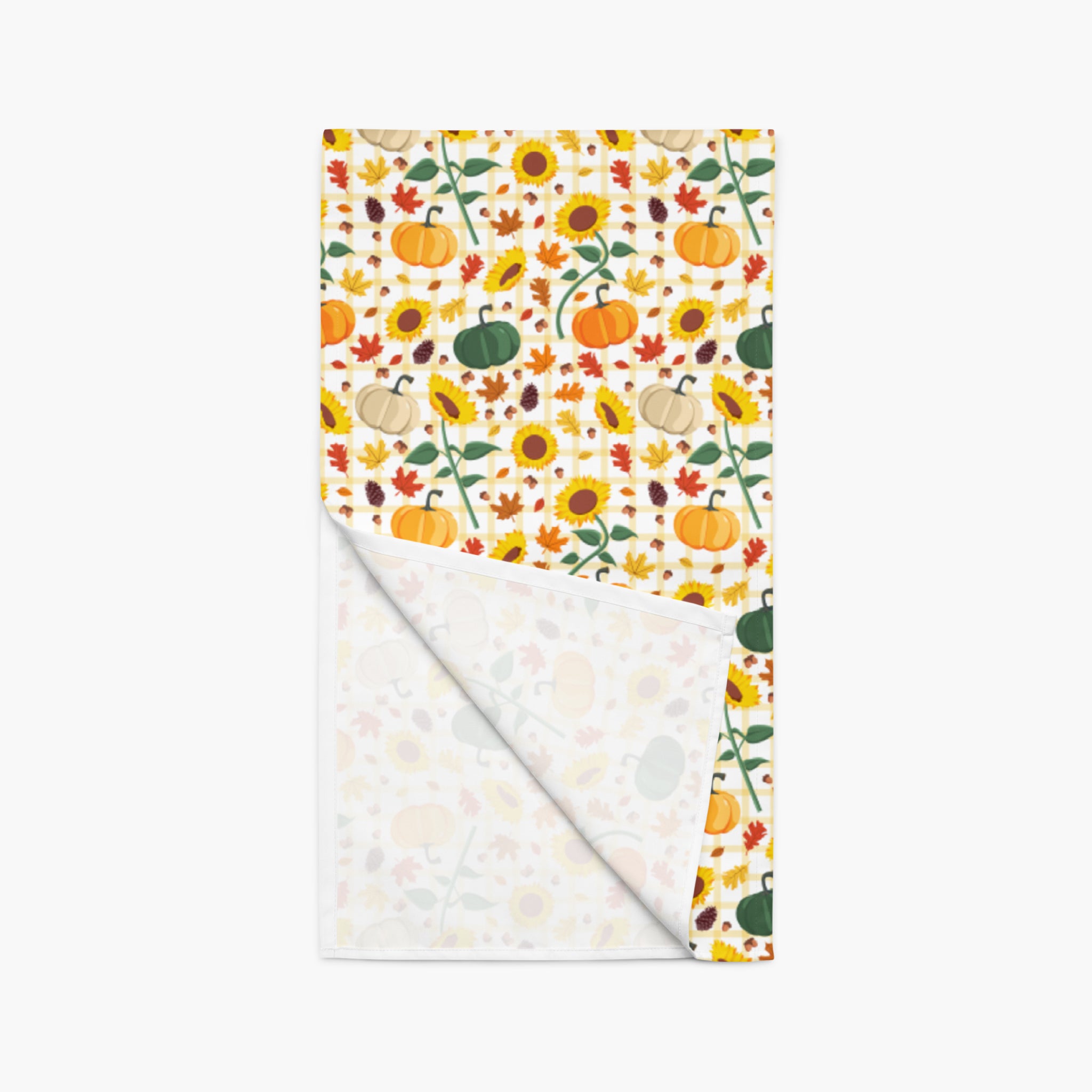 A folded table runner featuring a vibrant autumn pattern on one side and a solid white back side. The top side displays a dense pattern of pumpkins, sunflowers, maple leaves, acorns, and pinecones photographed on a light-grey studio background.