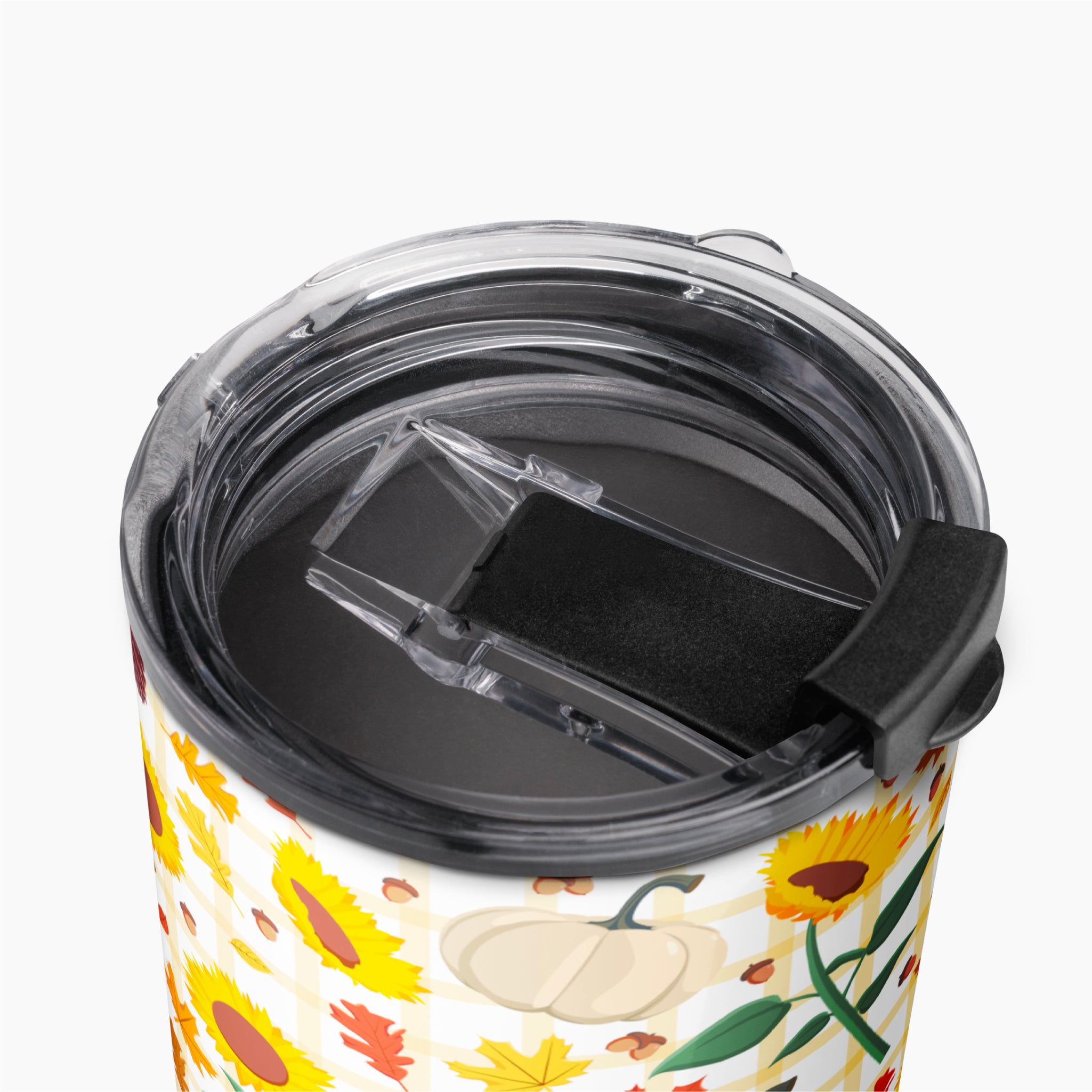 Close-up of a travel tumbler with a clear lid and black flip cover, featuring a fall harvest pattern with sunflowers, pumpkins, leaves, and acorns on a light-grey background.