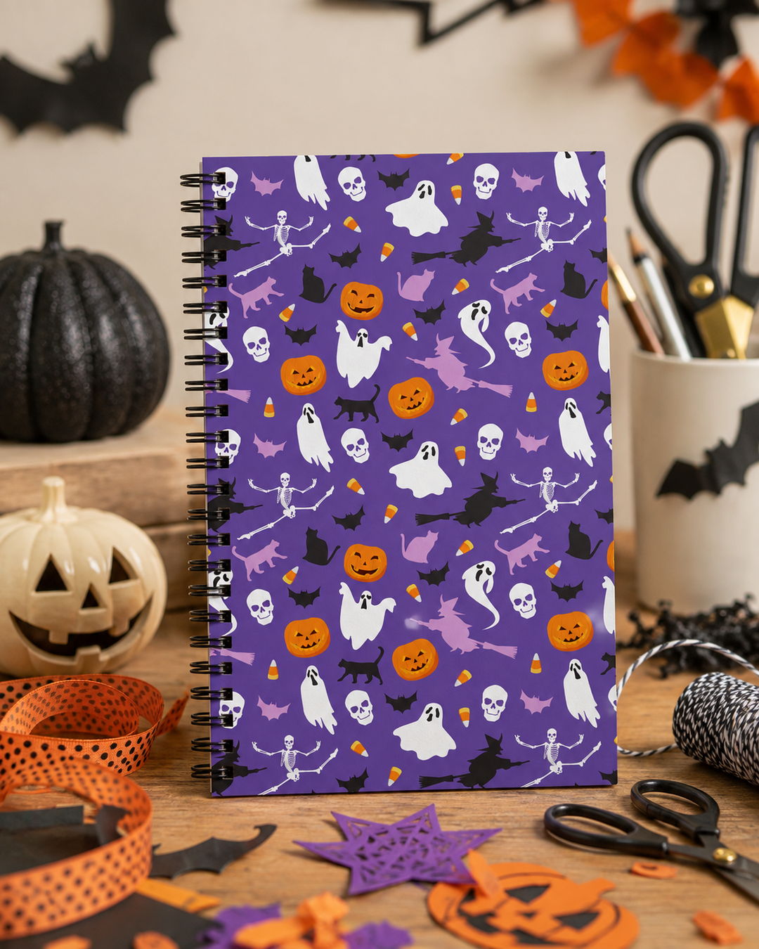 A spiral-bound notebook with a purple Halloween pattern featuring ghosts, pumpkins, bats, and skeletons, displayed on a wooden table surrounded by seasonal decorations.