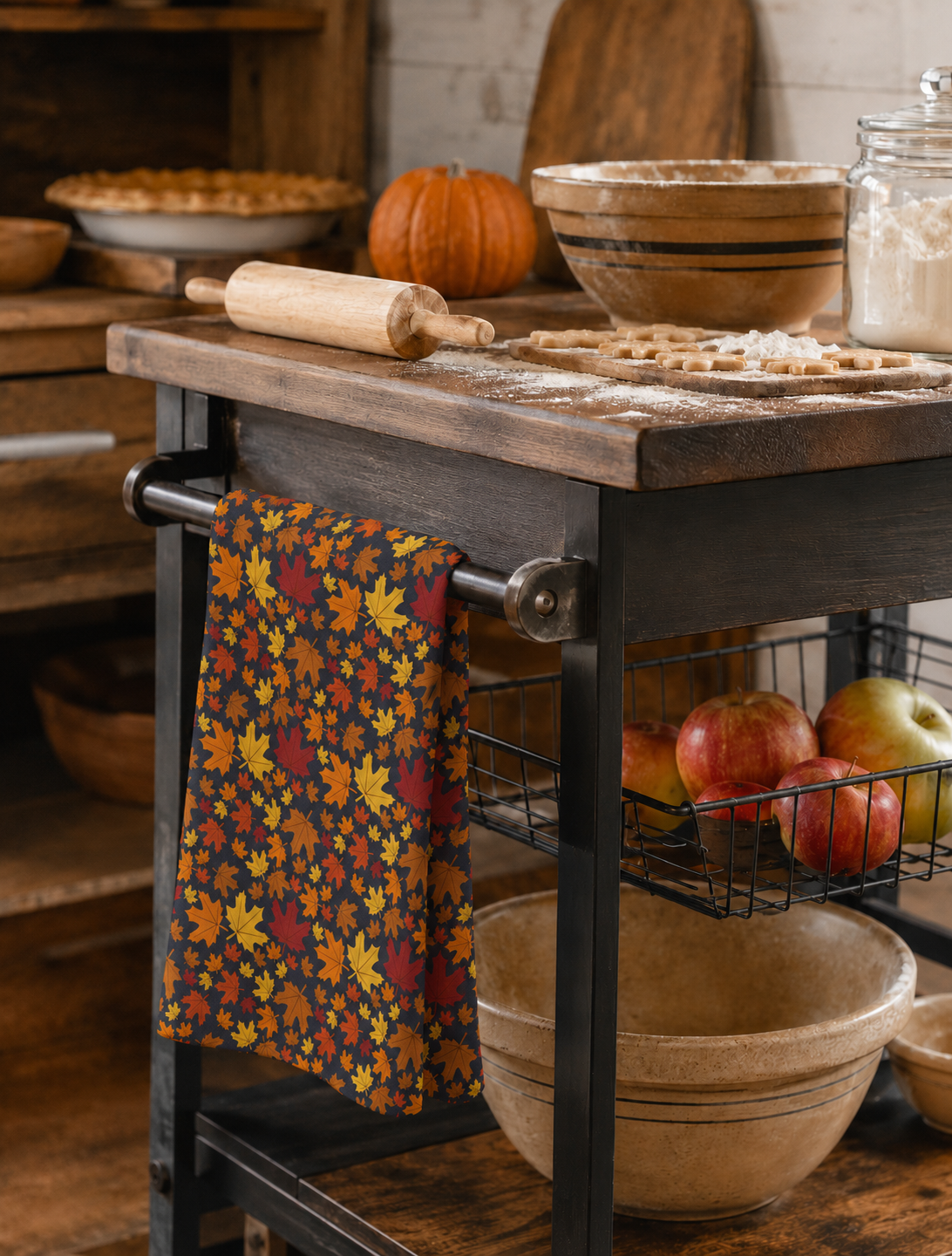 A kitchen scene featuring a rustic wooden rolling island cart with a dark metal frame. A colorful kitchen towel with an autumn leaf pattern hangs from the side bar.