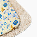 A close-up of a soft rectangular blanket with a cream sherpa backing and a printed top layer featuring blue menorahs, Star of David motifs, doves, and dreidel details on a light cream background.