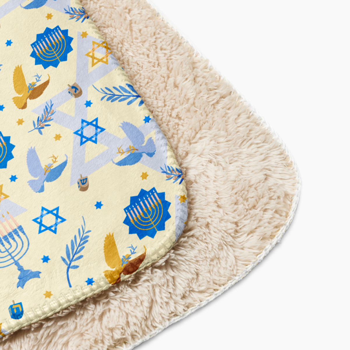 A close-up of a soft rectangular blanket with a cream sherpa backing and a printed top layer featuring blue menorahs, Star of David motifs, doves, and dreidel details on a light cream background.